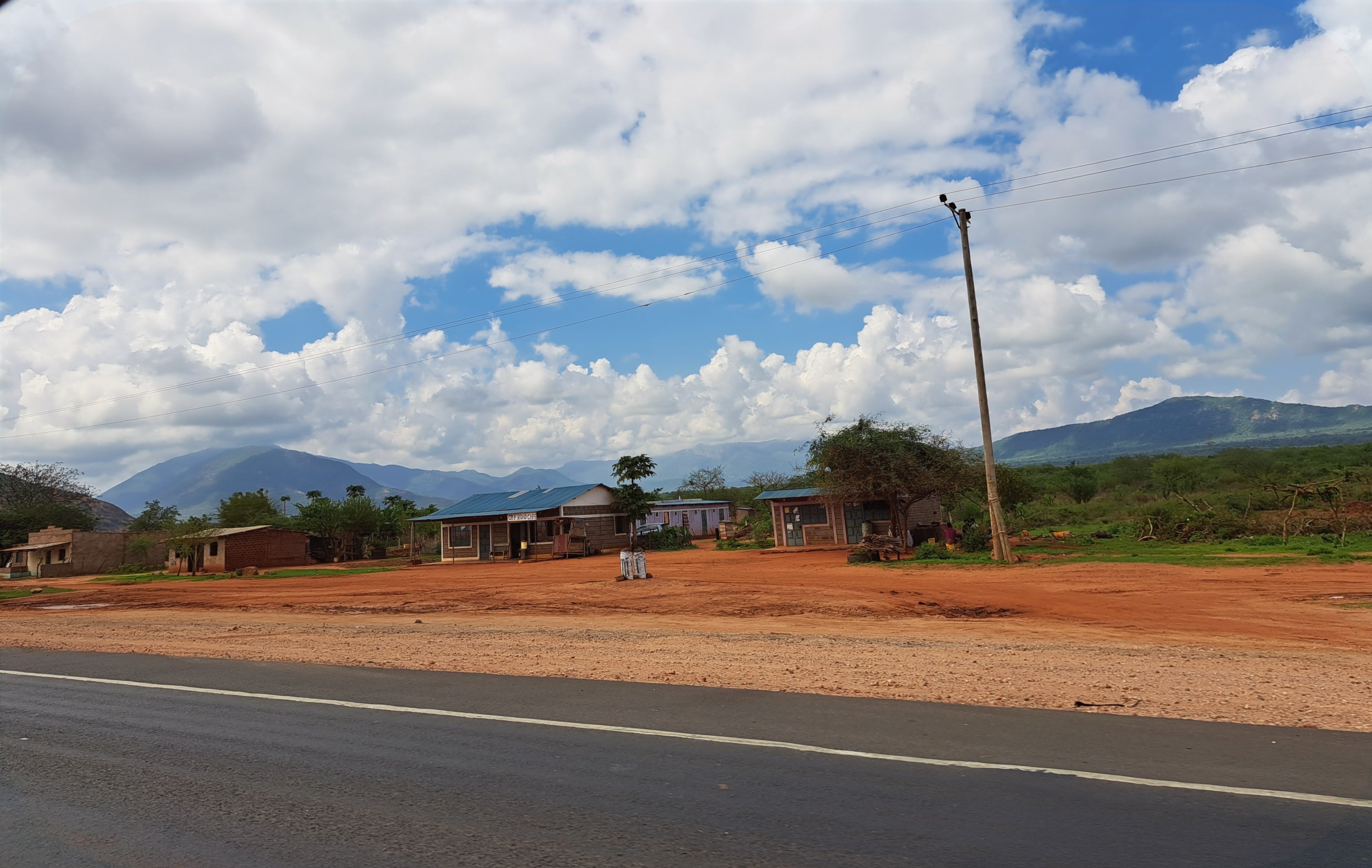Rural landscape with small roadside buildings, red earth, and distant mountains under bright clouds reflecting the everyday scenes across East Africa’s coffee‑growing regions