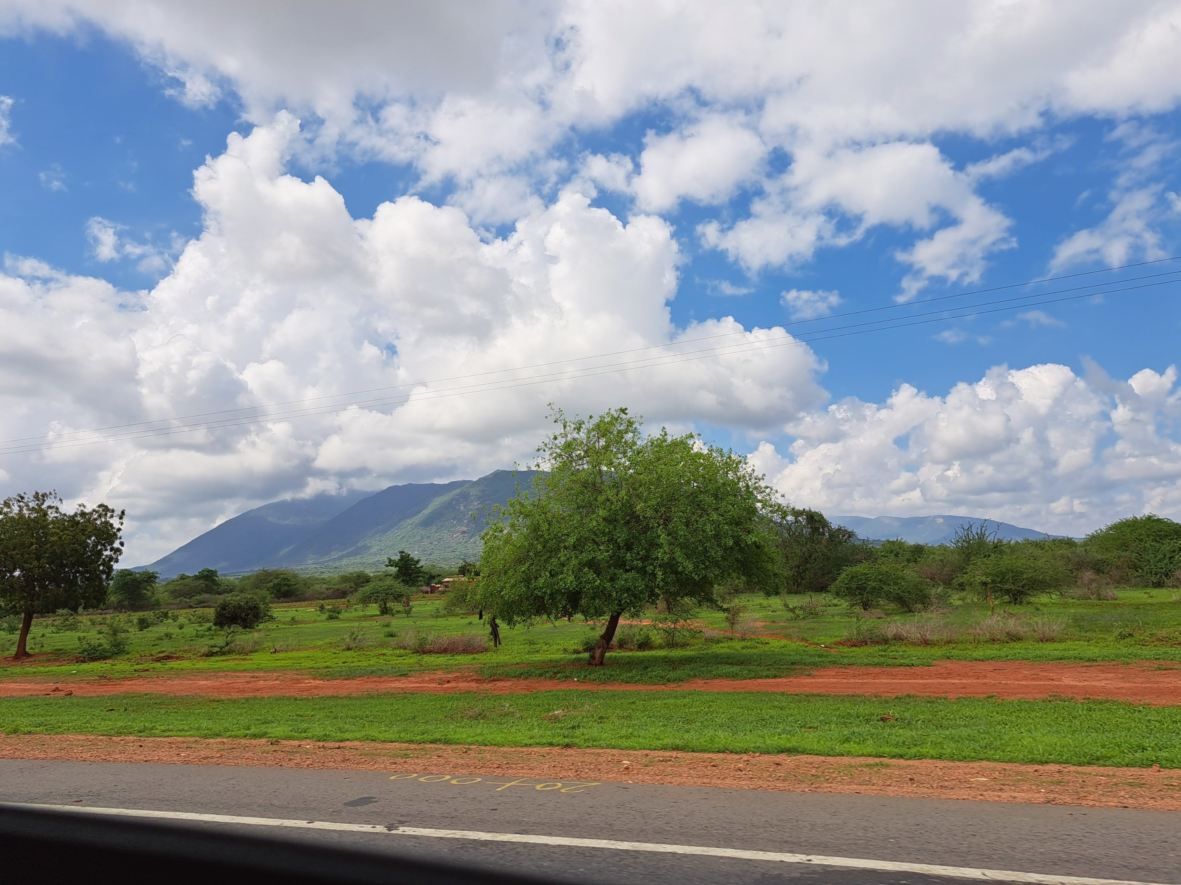 Scenic view of Kenya’s coffee highlands with green fields, red soil, and a mountain under bright blue skies representing the origin of Kenya Kafe Cup’s coffee